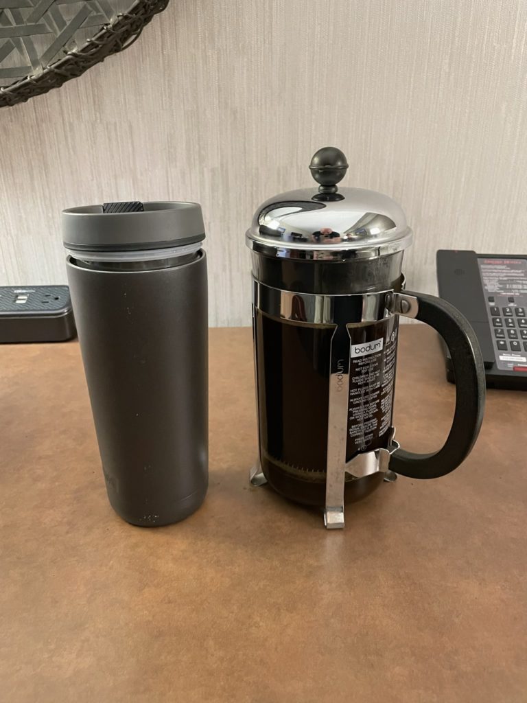A photo of a travel mug next to a French press full of coffee with the plunger depressed, ready to pour