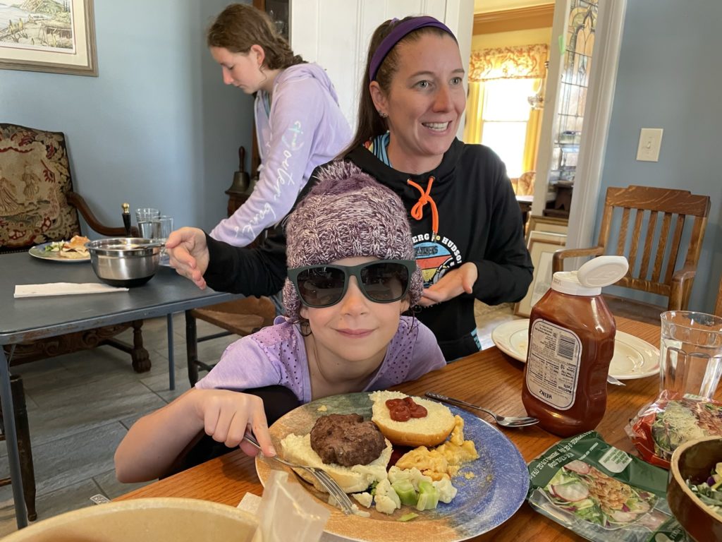 A photo of Ainsley wearing Kelsey's winter hat and sunglasses sitting at a table eating a hamburger and vegetables with Kelsey and Rayleigh in the background