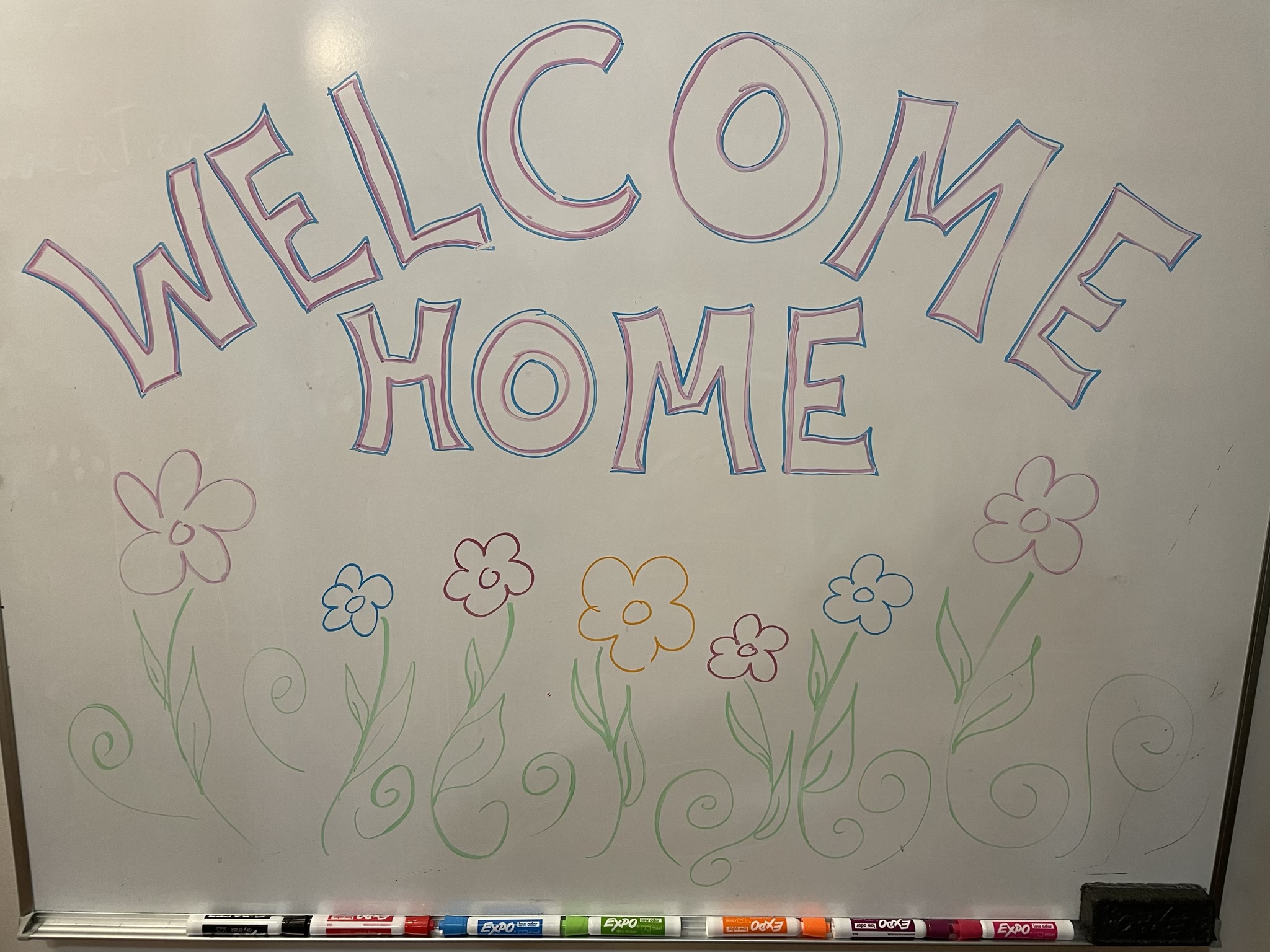 A photo of a whiteboard with the words "welcome home" written on it and drawings of spring flowers