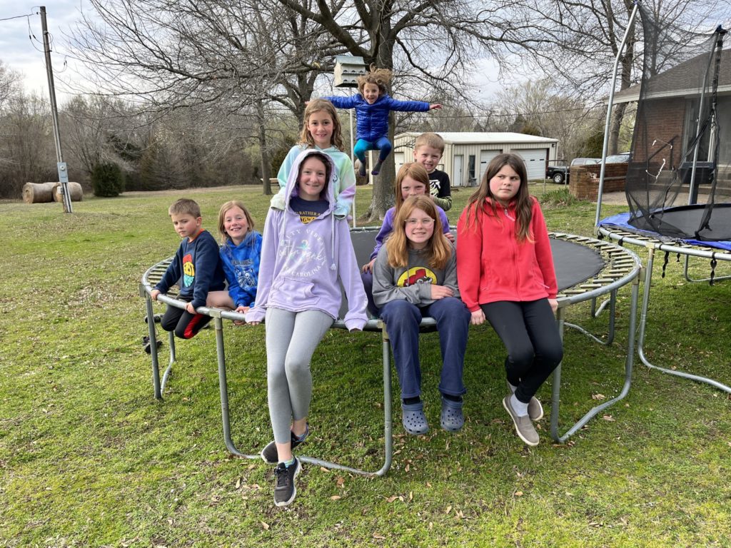 A photo of Oliver, Dillon, Rayleigh, Ainsley, Grayson, Kate, Nora, Peter, and Molly sitting on a trampoline. Grayson is bouncing and is midair in the photo.
