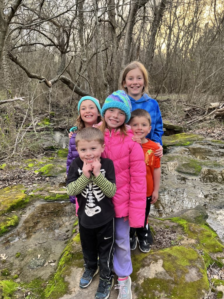 A photo of Peter, Ainsley, Nora, Oliver, and Dillon standing on a mossy rock by some standing water and trees
