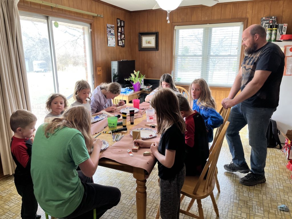 A photo of Peter, Kate, Grayson, Ainsley, Rayleigh, Molly, Nora, Oliver, and Dillon working on crafts at a table while Kirk provides direction