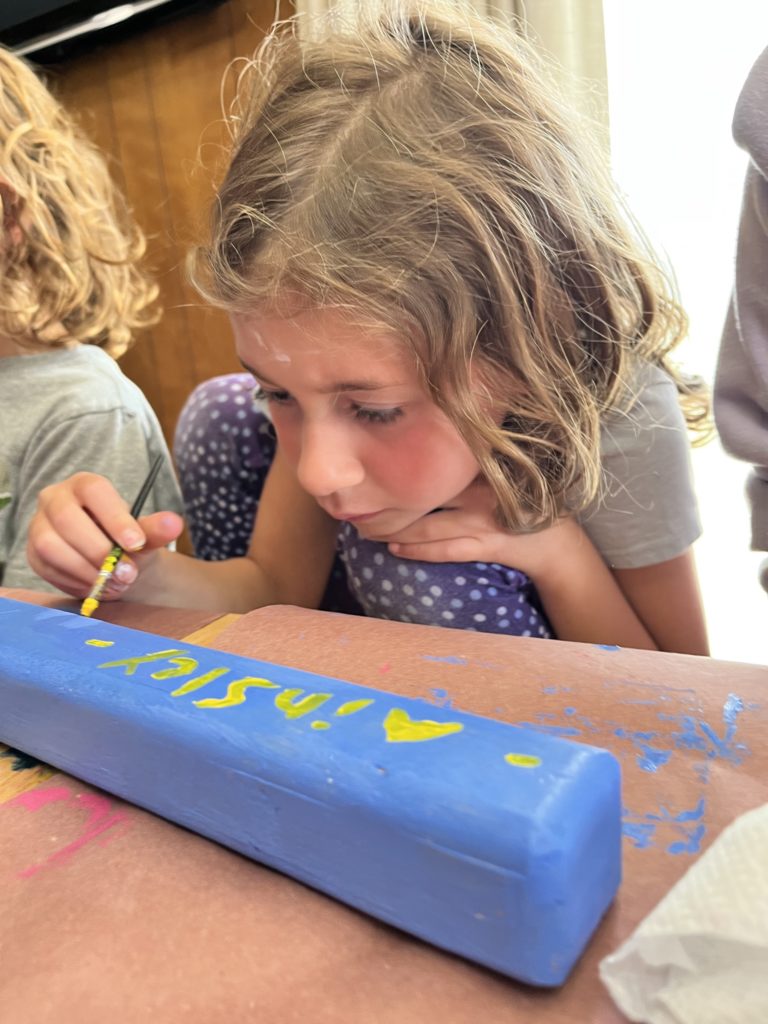 A photo of Ainsley sitting on a chair with her knees to her chest painting her name in yellow on a block of wood painted blue at a table