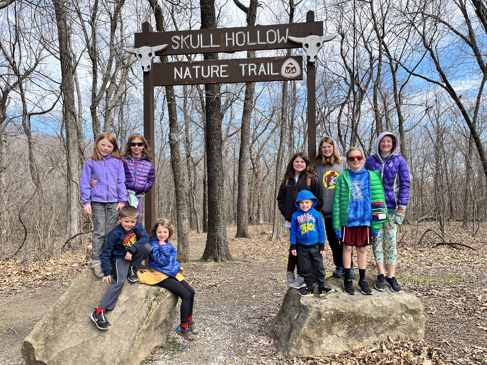 A photo of Nora, Ainsley, Peter, Grayson, Molly, Oliver, Kate, Dillon, and Rayleigh standing on rocks wearing coats and sweatshirts outside under a sign that says "Skull Hollow Nature Trail" with a picture of a longhorn cow skull