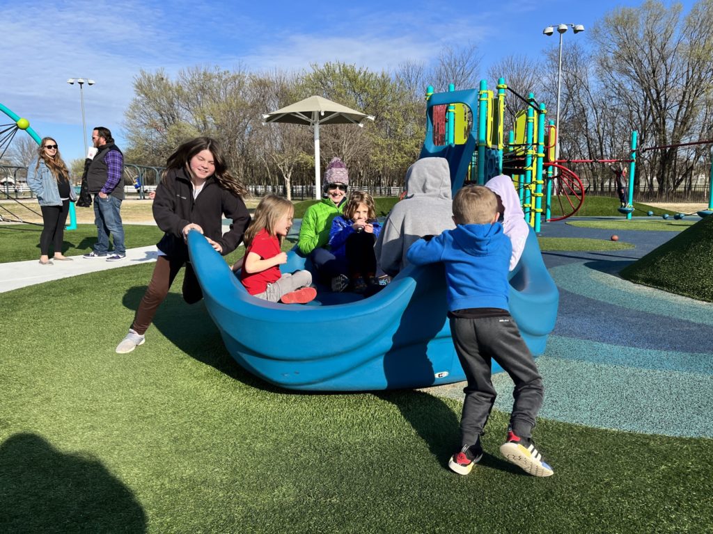 A photo of Molly and Oliver spinning a piece of playground equipment with Kelsey, Grayson, Kate, Rayleigh, and another child in it