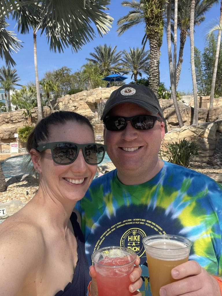 A photo of Kelsey and Kevin in sunglasses with colorful beverages in plastic cups on the pool deck with palm trees in the background