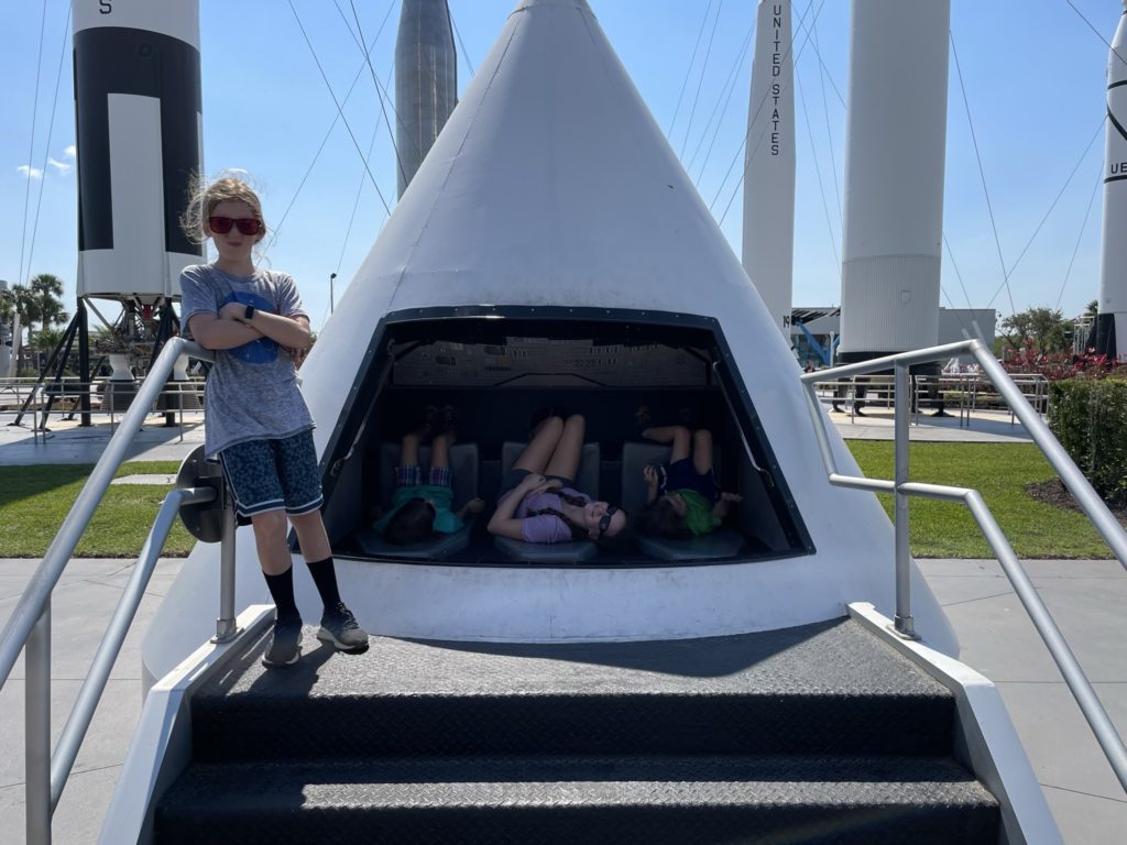 A photo of Dillon making a frowny face while Ainsley, Rayleigh, and Grayson sit in the only three seats aboard a model of a spaceship