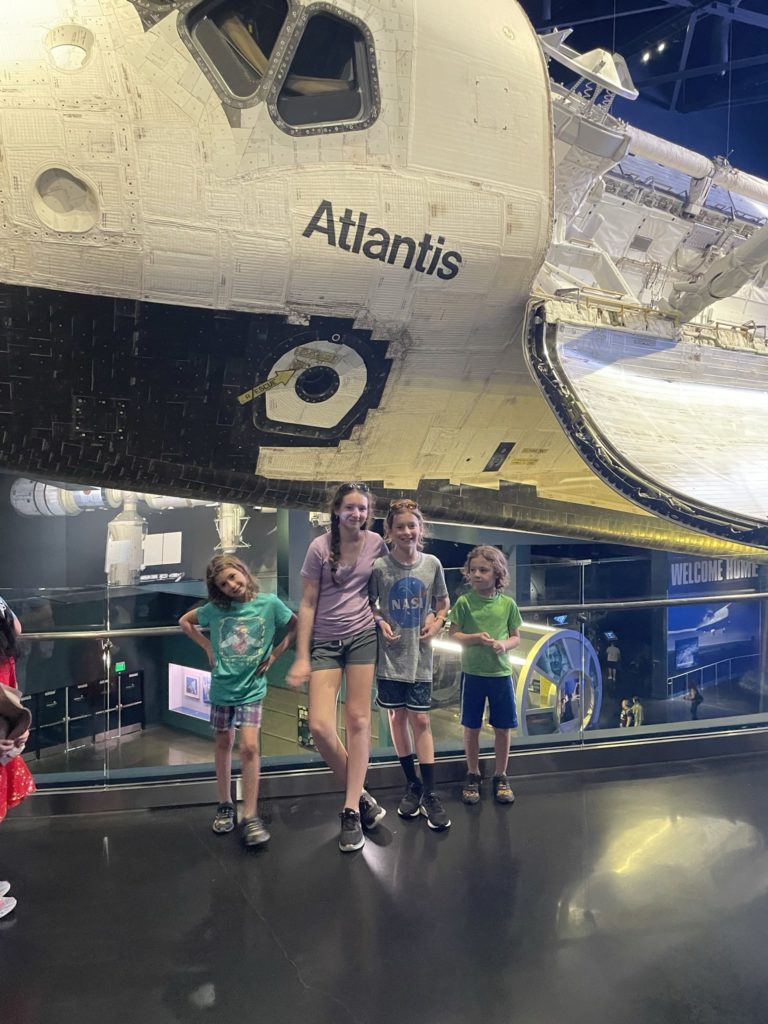 A photo of Ainsley, Rayleigh, Dillon, and Grayson standing in front of the Atlantis space shuttle, which is suspended from the ceiling