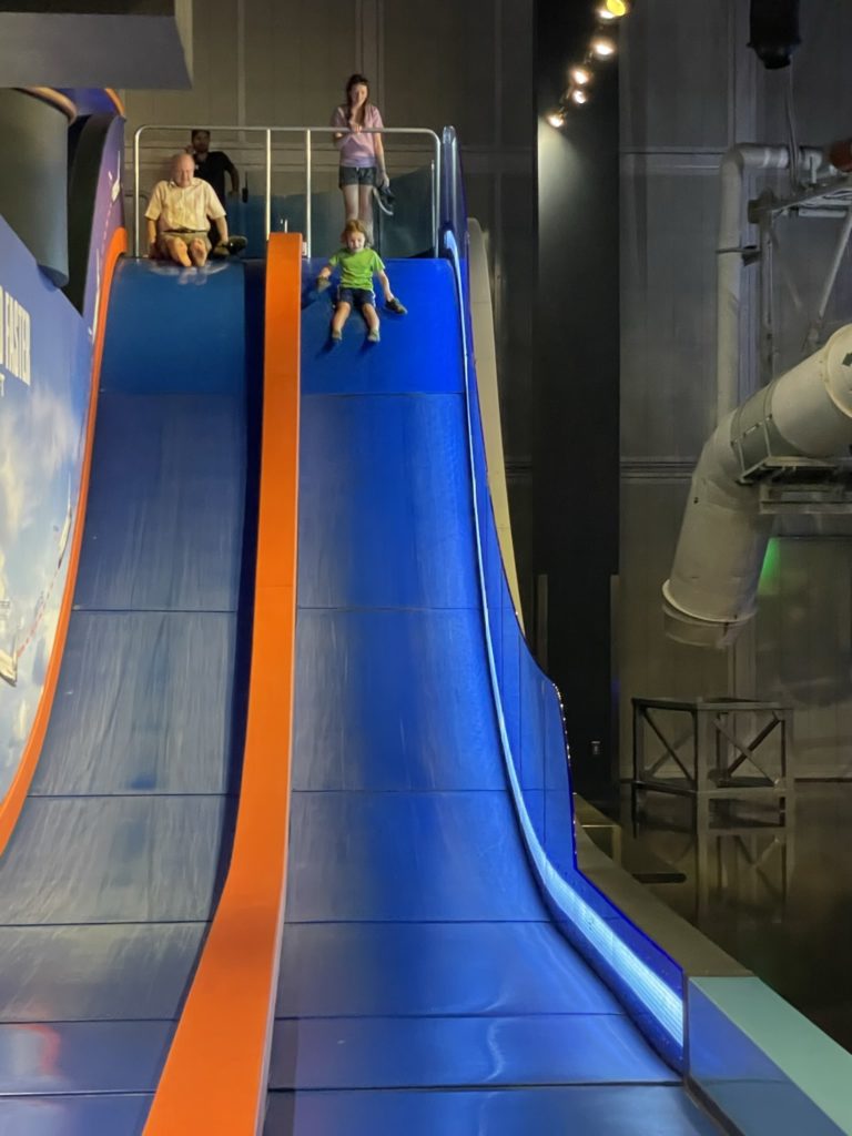 A photo of Grayson going down a large blue slide while Rayleigh waits her turn