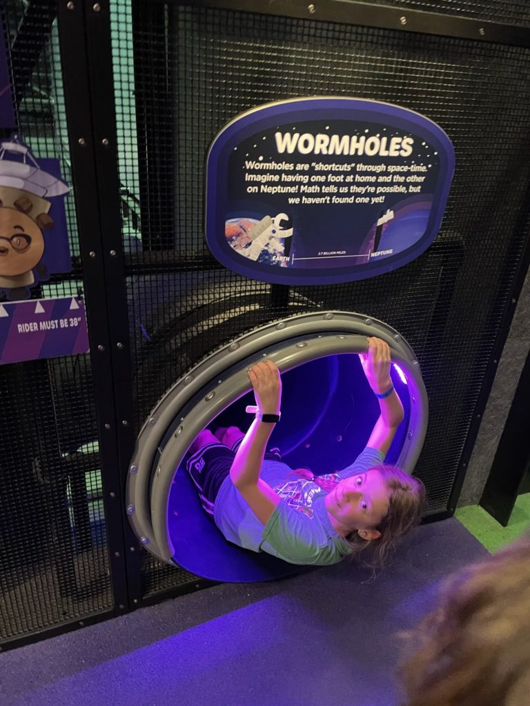 A photo of Dillon climbing into a tunnel that is under a sign that says "Wormholes" and has some information about wormholes on it