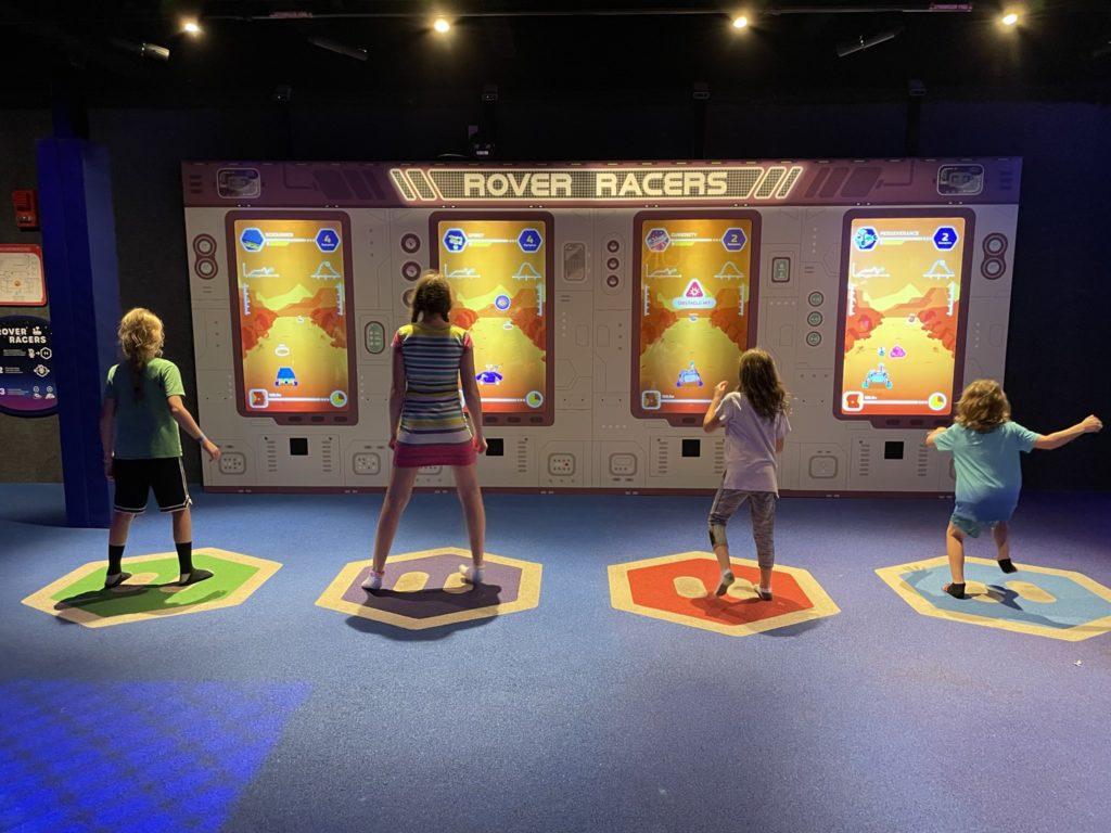 A photo of Dillon, Rayleigh, Ainsley, and Grayson playing a video game that is controlled by the movement of their bodies called "Rover Racers"