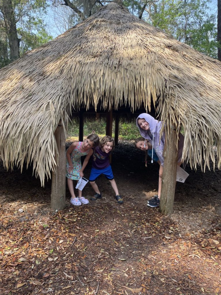 A photo of Ainsley, Grayson, Dillon, and Rayleigh inside of a low thatched roof shelter with no walls