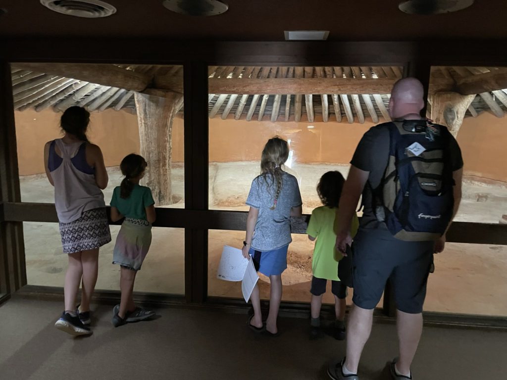 A photo of Rayleigh, Ainsley, Dillon, Grayson, and Kevin inside one of the mounds, which contains a packed dirt floor with straight clay walls and a roof supported by wood beams