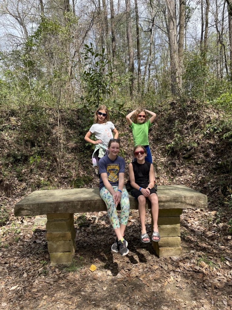 A photo of Ainsley, Grayson, Rayleigh, and Dillon sitting on a large bench in the woods