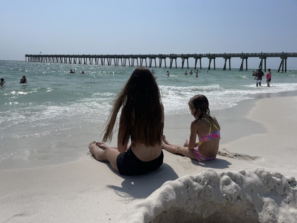 A photo of Kahlan and Ainsley sitting on the beach with a pier in the background