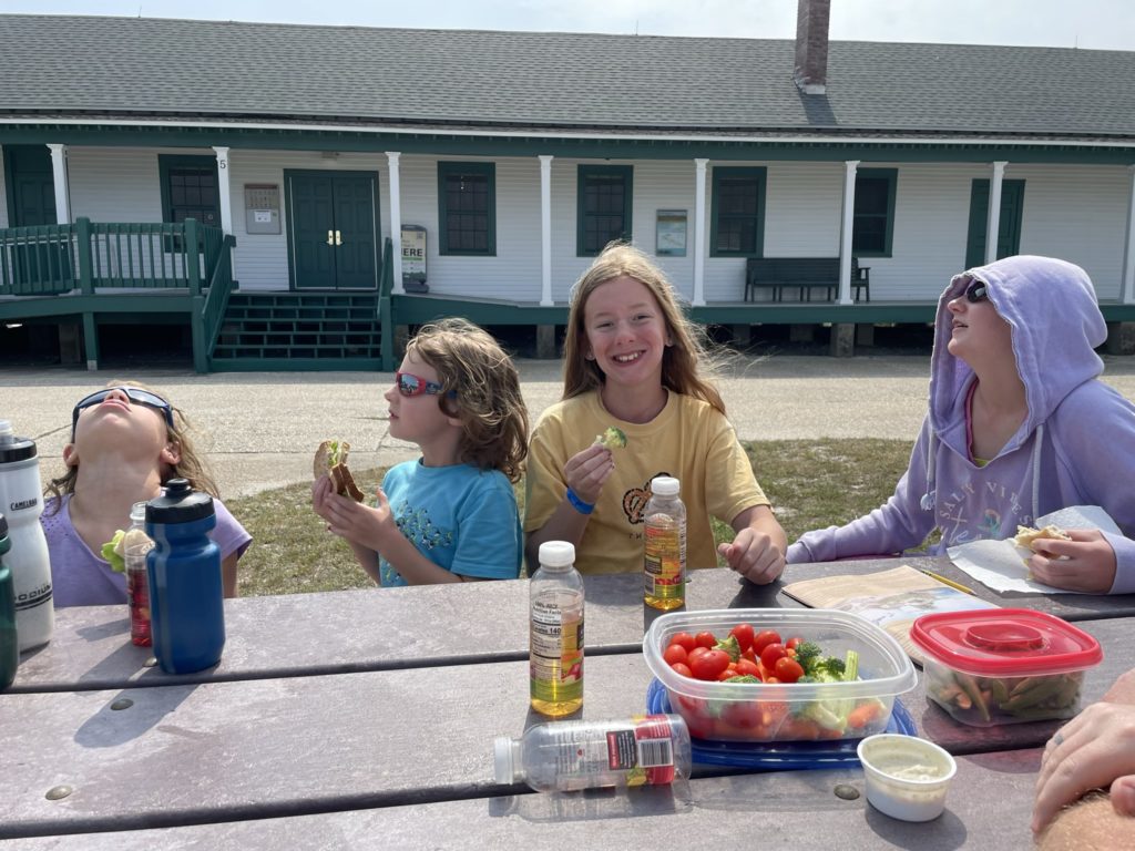 A photo of Ainsley, Grayson, Dillon, and Rayleigh sitting at a picnic table eating lunch outside with the national park visitor's center in the background