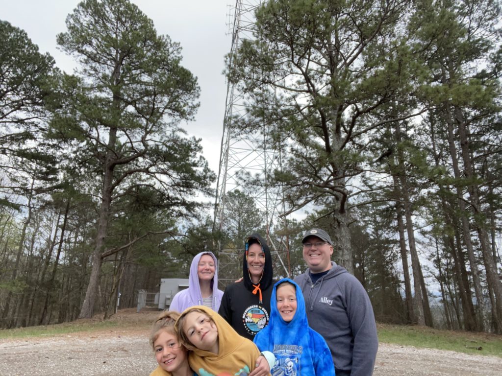 A photo of Rayleigh, Kelsey, Kevin, Ainsley, Grayson, and Dillon standing on top Mount Woodall, the tallest point in the state of Mississippi, with trees and a communications tower in the background