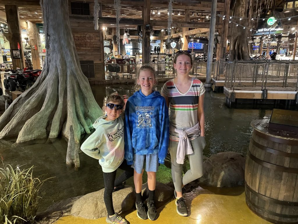 A photo of Ainsley, Dillon, and Rayleigh standing in front of an indoor pond and fake tree with retail space in the background