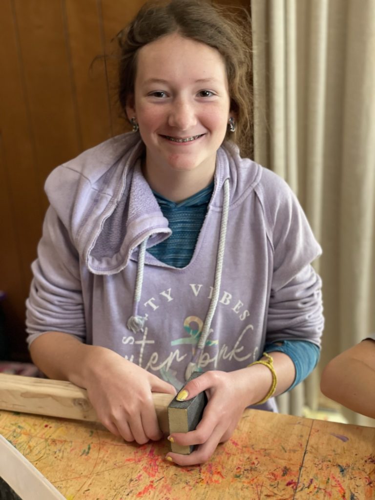 A photo of Rayleigh wearing a hoodie and smiling at the camera while she sands a block of wood at a table