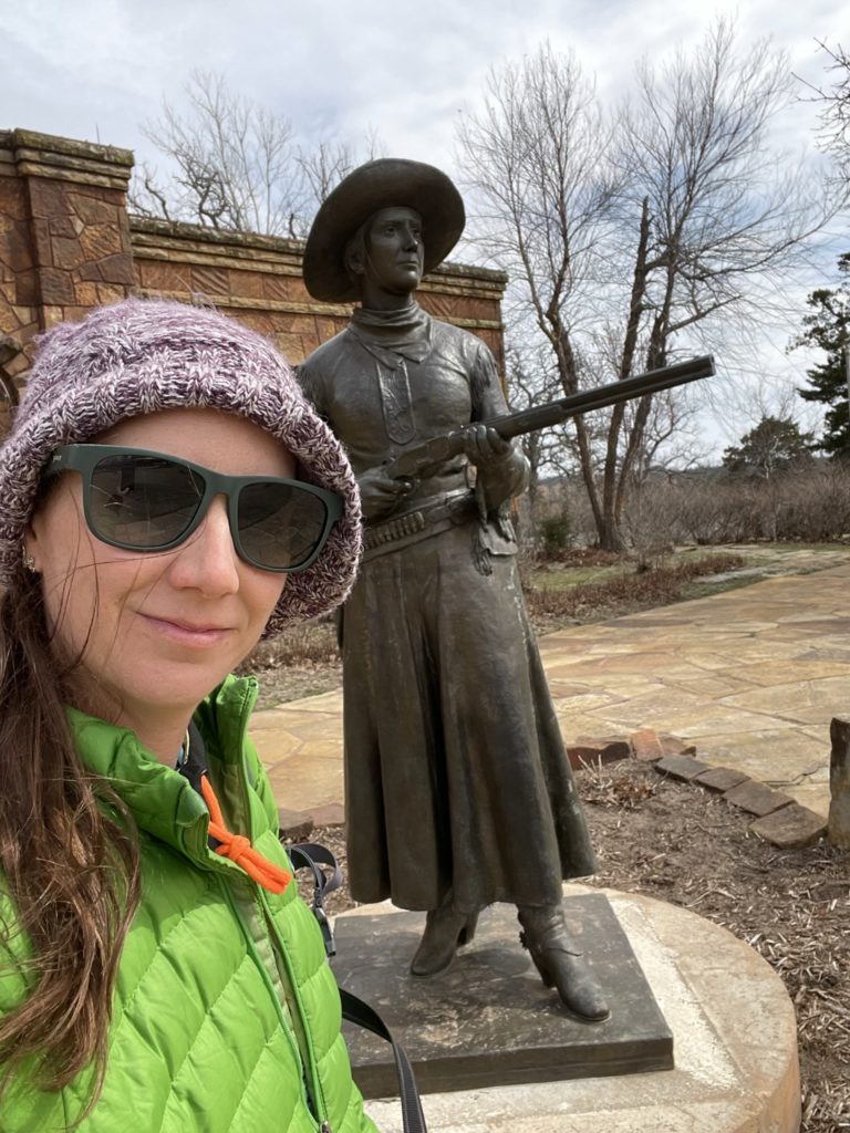 A photo of Kelsey standing next to a statue of Belle Starr, who is wearing a wide brimmed hat and pointing a long gun