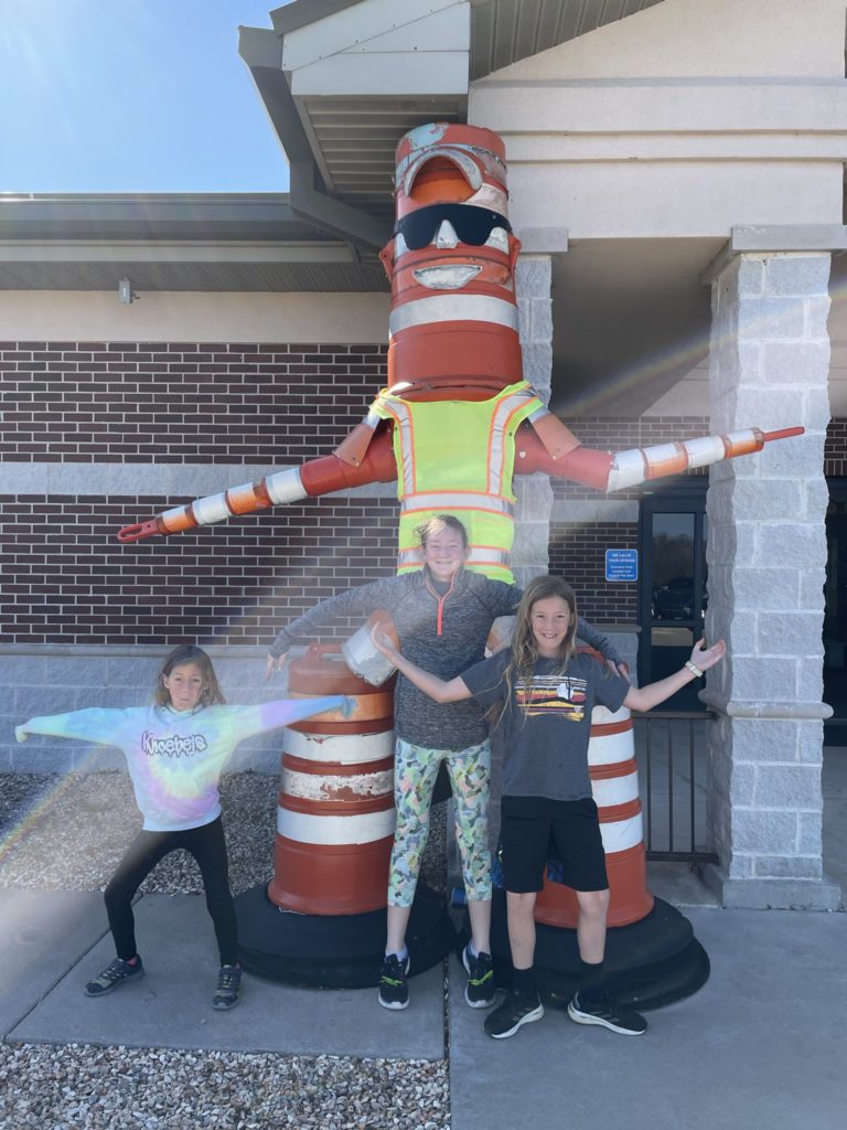 A photo of Ainsley, Rayleigh, and Dillon posing next to a 10 foot tall "man" made out of construction cones and a reflective vest