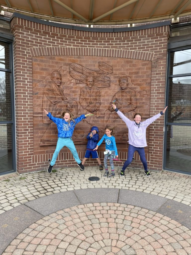 A photo of Dillon, Grayson, Ainsley, and Rayleigh jumping and making silly poses in front of a brick mural to the Wright brothers