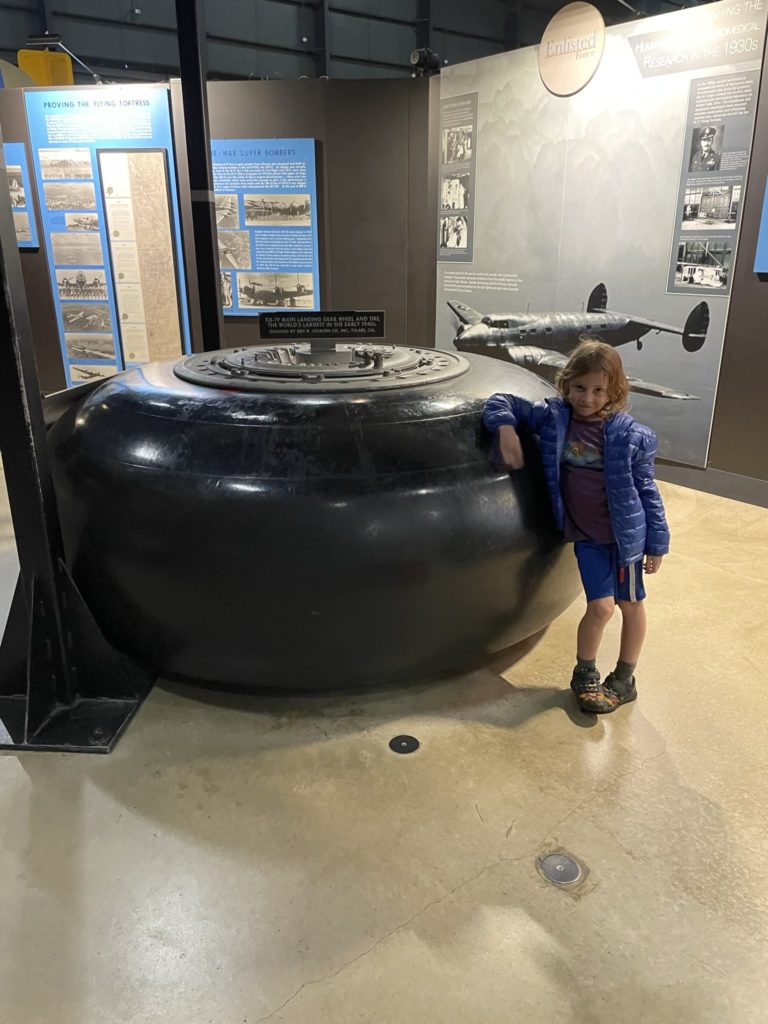 A photo of Grayson standing in front of an airplane tire laying on its side, which is nearly as tall as he is. The sign on top reads "XB-19 main landing gear wheel and tire, the world's largest in the early 1940s"