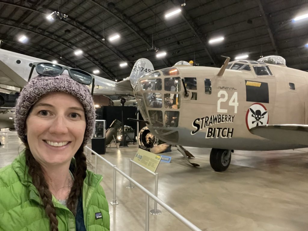 A photo of Kelsey standing in front of a plane that says "Strawberry Bitch" on the side