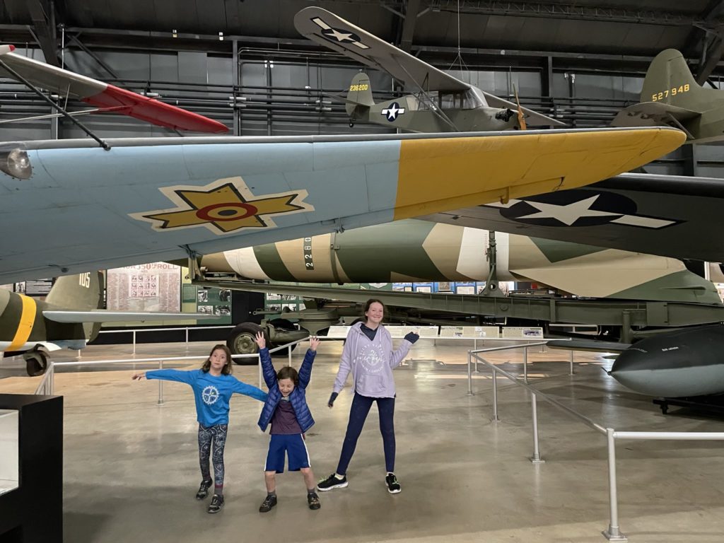 A photo of Ainsley, Grayson, and Rayleigh posing under two overlapping aircraft wings