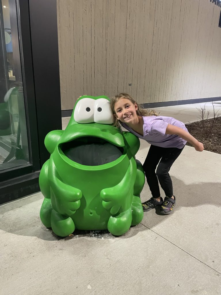 A photo of Ainsley standing next to a statue of a large green frog that's nearly as tall as she is outside of an Interstate rest area
