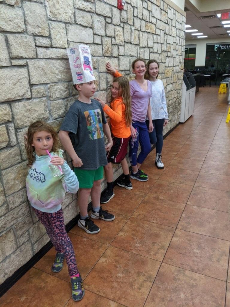 A photo of Ainsley, Milo, Dillon, Rayleigh, and Myna posing next to a stone wall in a restaurant. Milo has a paper bag on his head like a hat and Dillon is pretending to climb the wall.